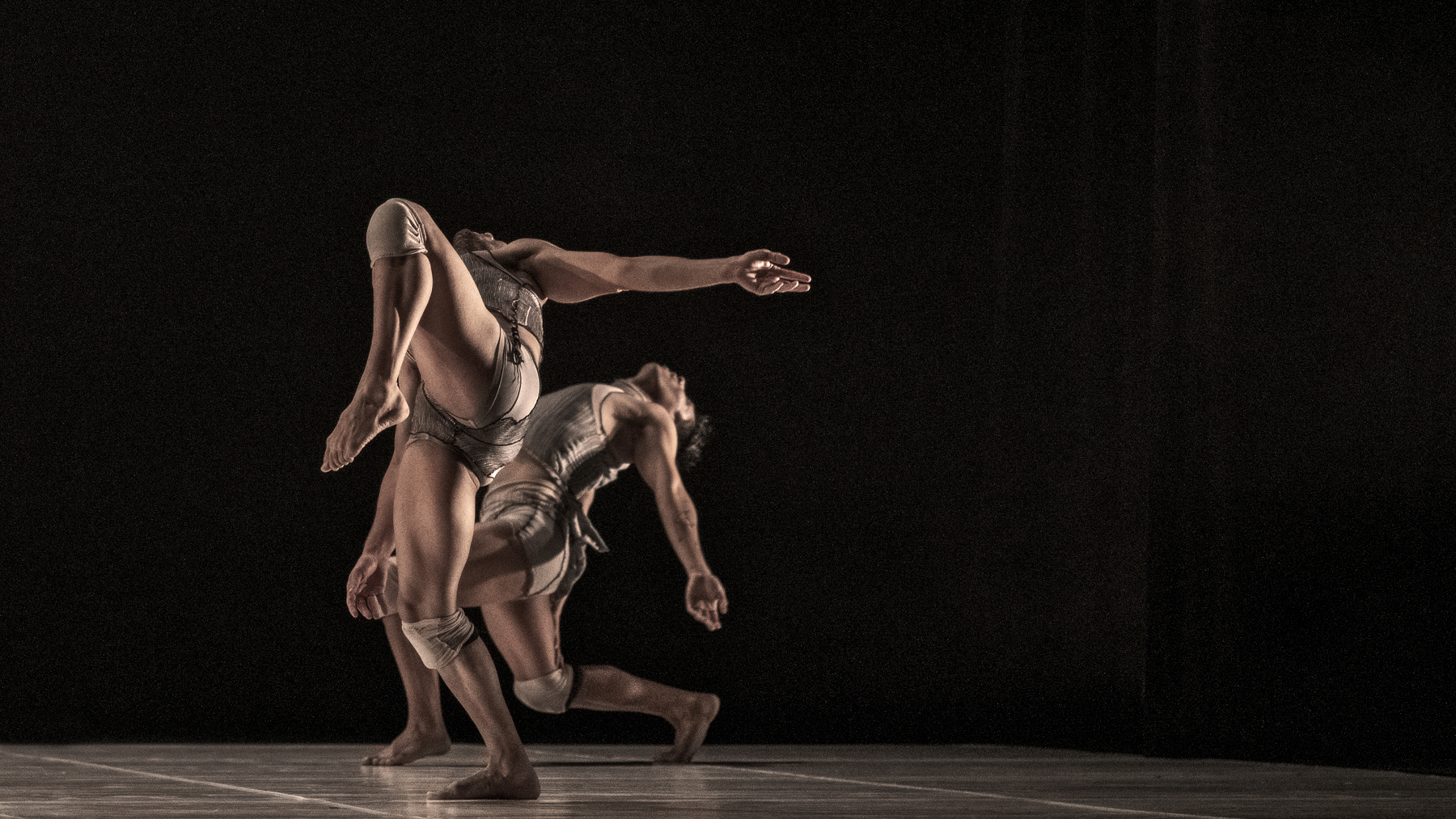 Two dancers in gray costumes leap across a dark stage, one arm extended forward as their bodies arc through the motion.