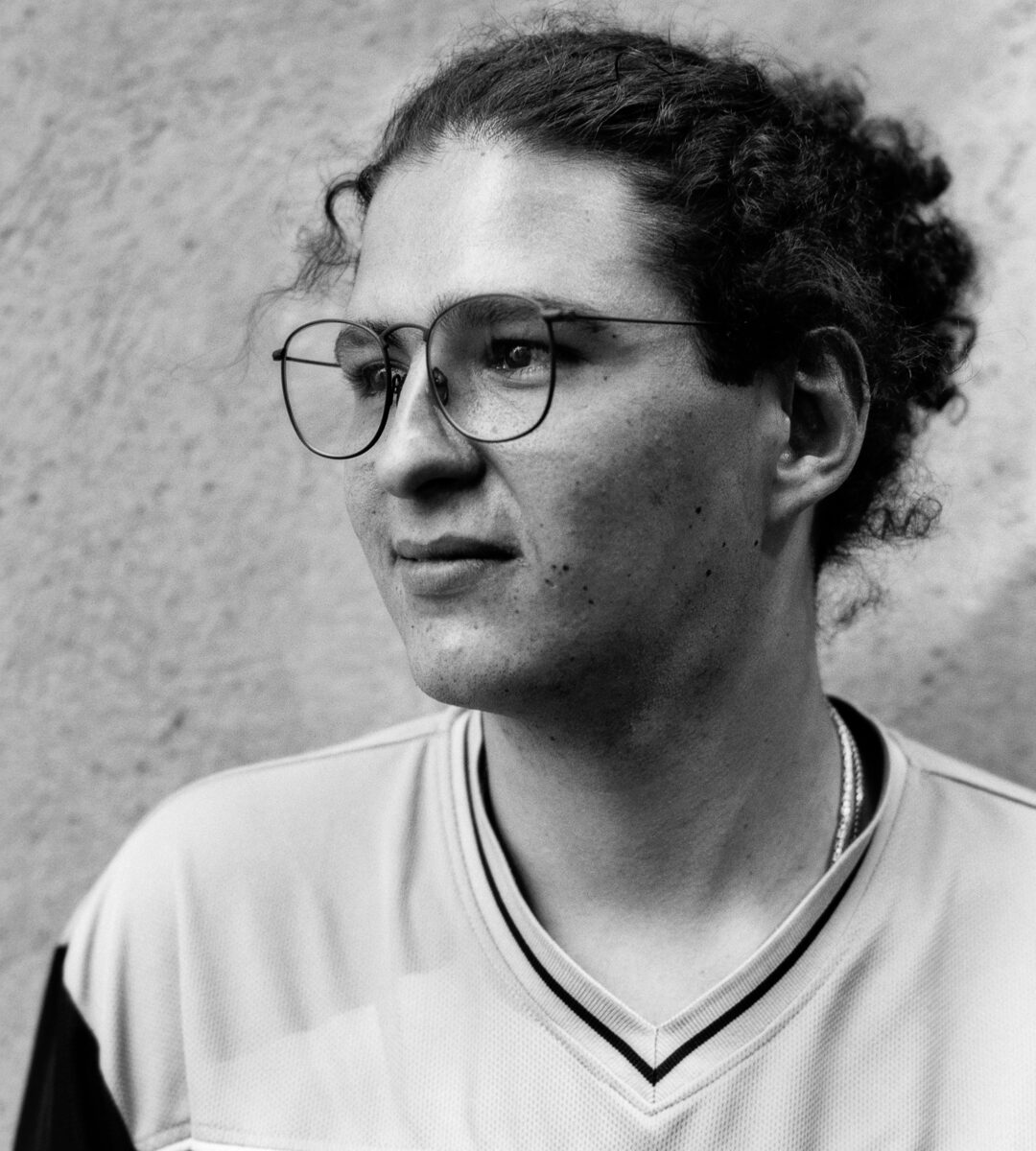 Black-and-white portrait of a young man with round glasses looking to the left, wearing a V-neck top.