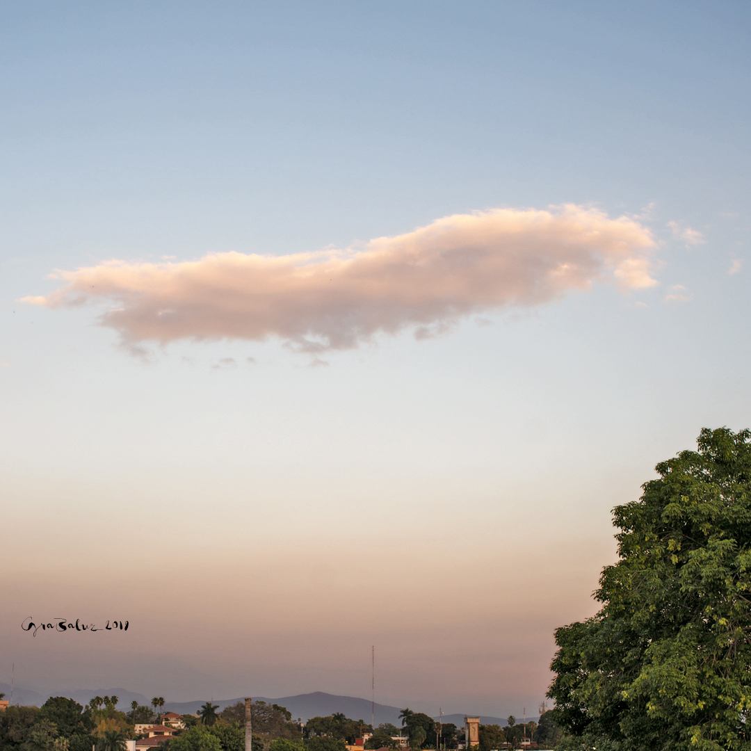 Pastel sunset sky over a small town with a single elongated cloud, distant hills, and a large tree on the right.